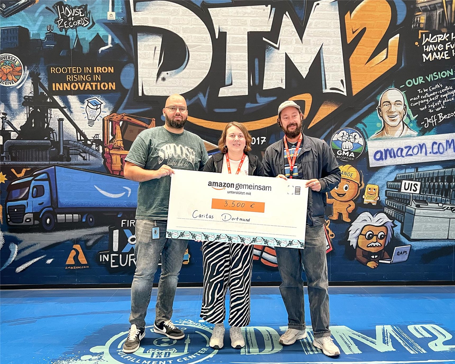 Links im Bild sieht man Christos Mazarakis von Amazon, der Kristina Sobiech von youngcaritas und Jan Hünicke von der Bbdachlosenseelsorge einen Scheck über 3500 € überreicht. 
