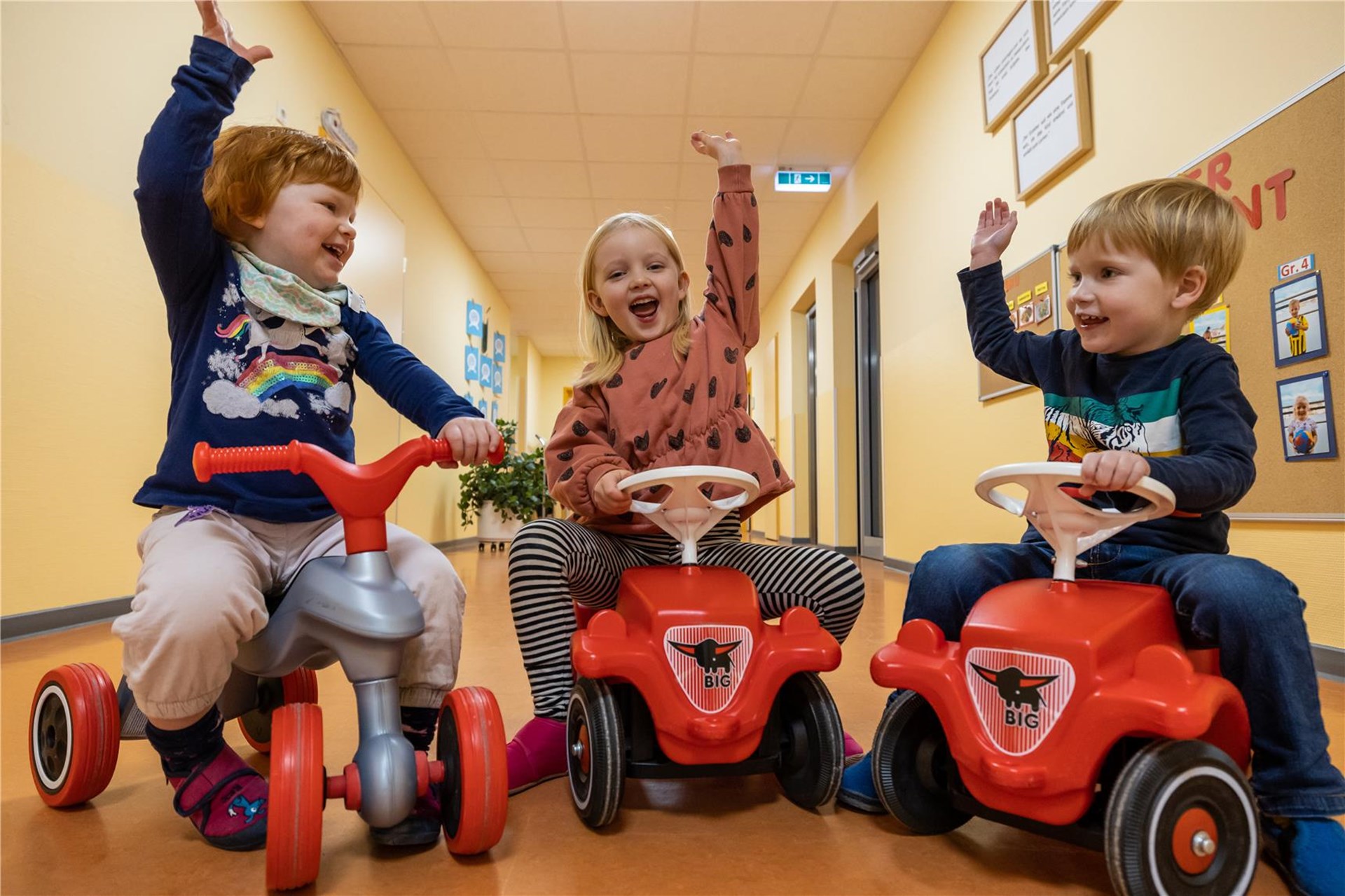 Drei Kinder sitzen auf Bobbycars und halten jubelnd die Hände in die Luft.