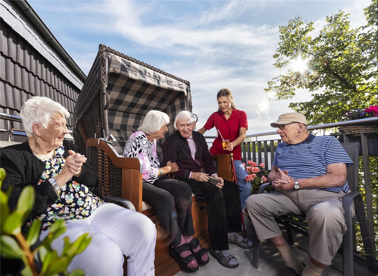 Senior:innen sitzen bei strahlendem Sonnenschein gemeinsam auf einer Terrasse, zwei davon in einem Starndkorb. Eine Pflegekraft reicht ein Getränk.