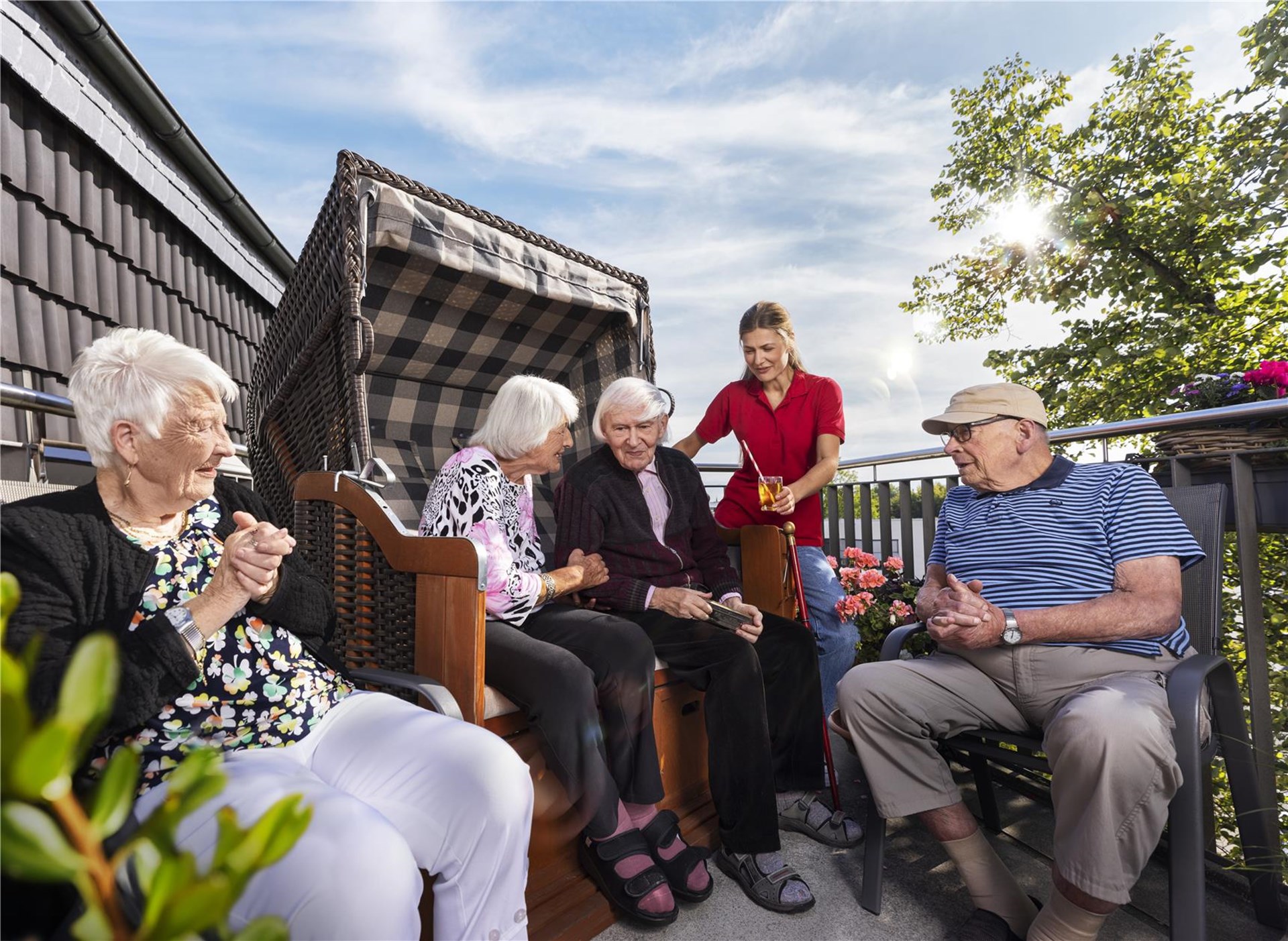 Senior:innen sitzen bei strahlendem Sonnenschein gemeinsam auf einer Terrasse, zwei davon in einem Starndkorb. Eine Pflegekraft reicht ein Getränk.
