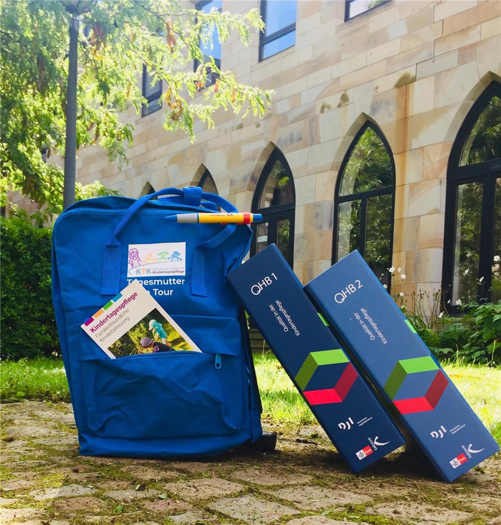 Ein Rucksak und zwei Ordner mit der Aufschrift QHB stehen auf einer Wiese im Propsteihof.