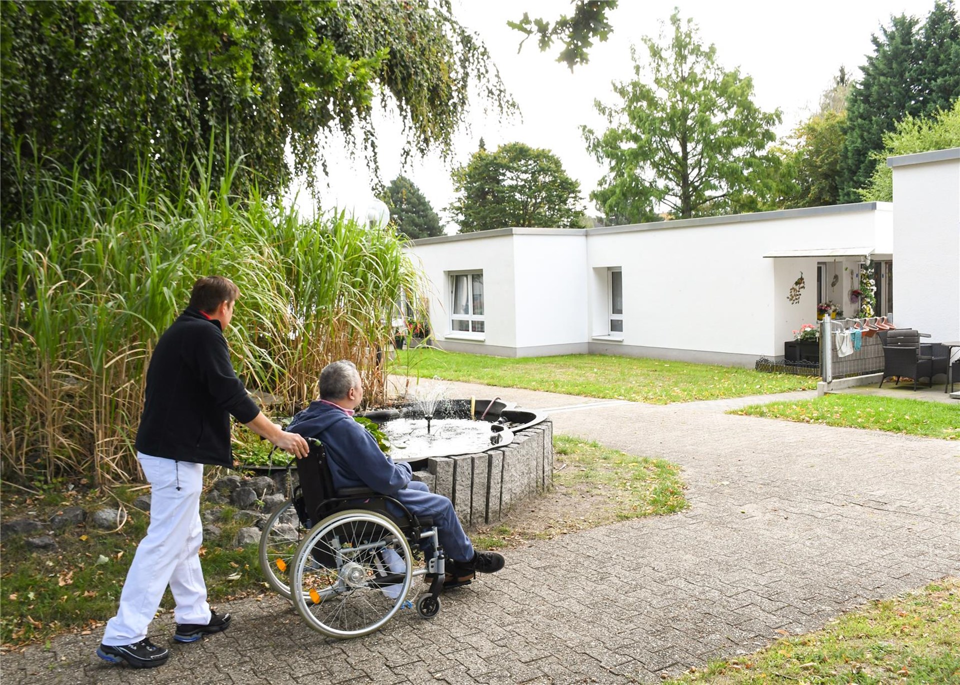 Mann fährt anderen Mann im Rollstuhl durch einen Garten.
